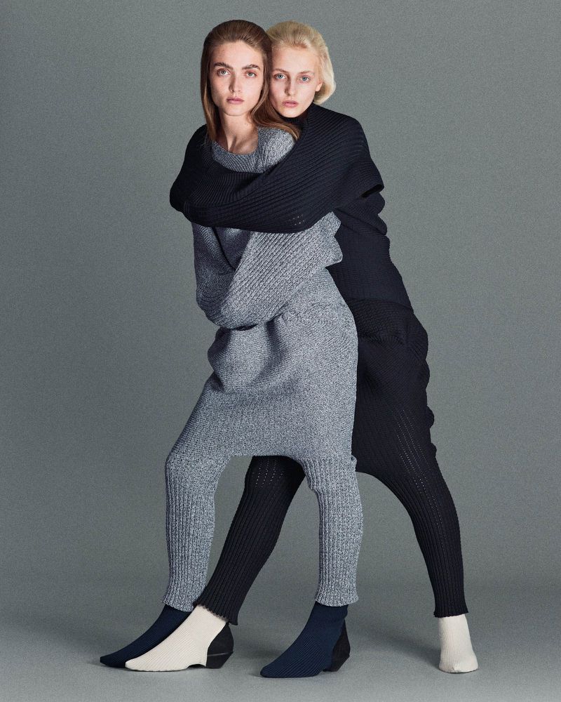 Two female models embracing closely, wearing textured grey and black knitwear against a neutral grey studio background