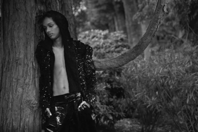 Model leaning against a tree trunk in a wooded area, wearing a dark sequined jacket and trousers