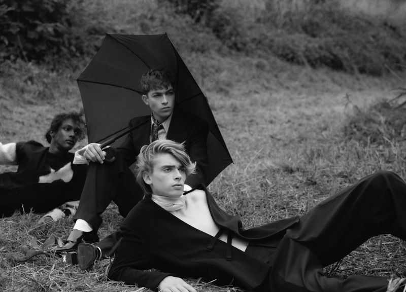 Three male models in dark tailoring posing outdoors on grassy ground with a large black umbrella