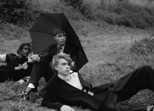 Three male models in dark tailoring posing outdoors on grassy ground with a large black umbrella