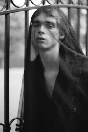 Model wearing a dark veil and coat standing behind an ornate wrought iron fence in a black and white portrait.