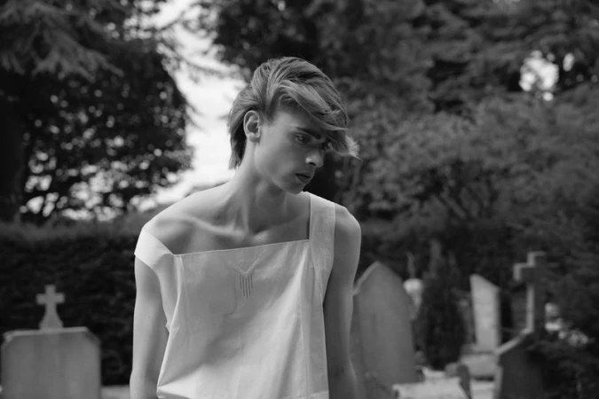 Monochrome portrait of a young male model wearing an asymmetrical white top outdoors among headstones.