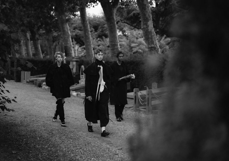 Three models walking along a path lined with tall trees in a black and white editorial fashion photograph.