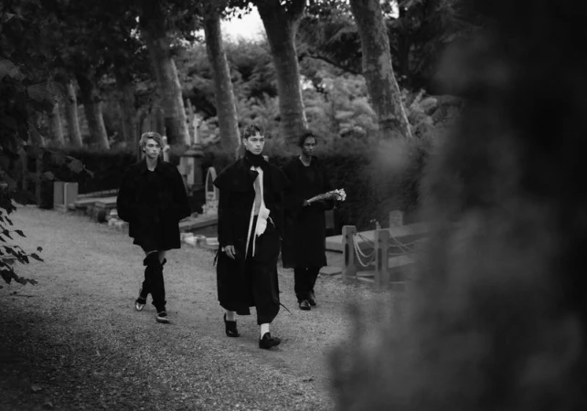 Three models walking along a path lined with tall trees in a black and white editorial fashion photograph.