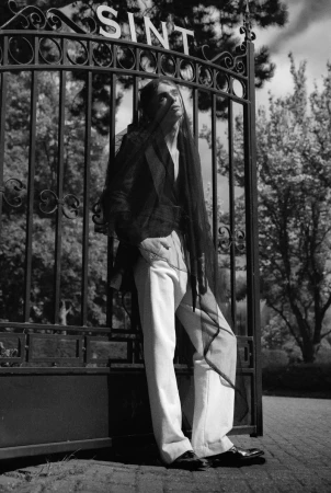 Monochrome fashion editorial image of a model leaning against an ornate iron gate with the word SINT visible above.