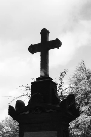 Monochrome low-angle shot of a dark stone cross monument against a cloudy sky with bare tree branches