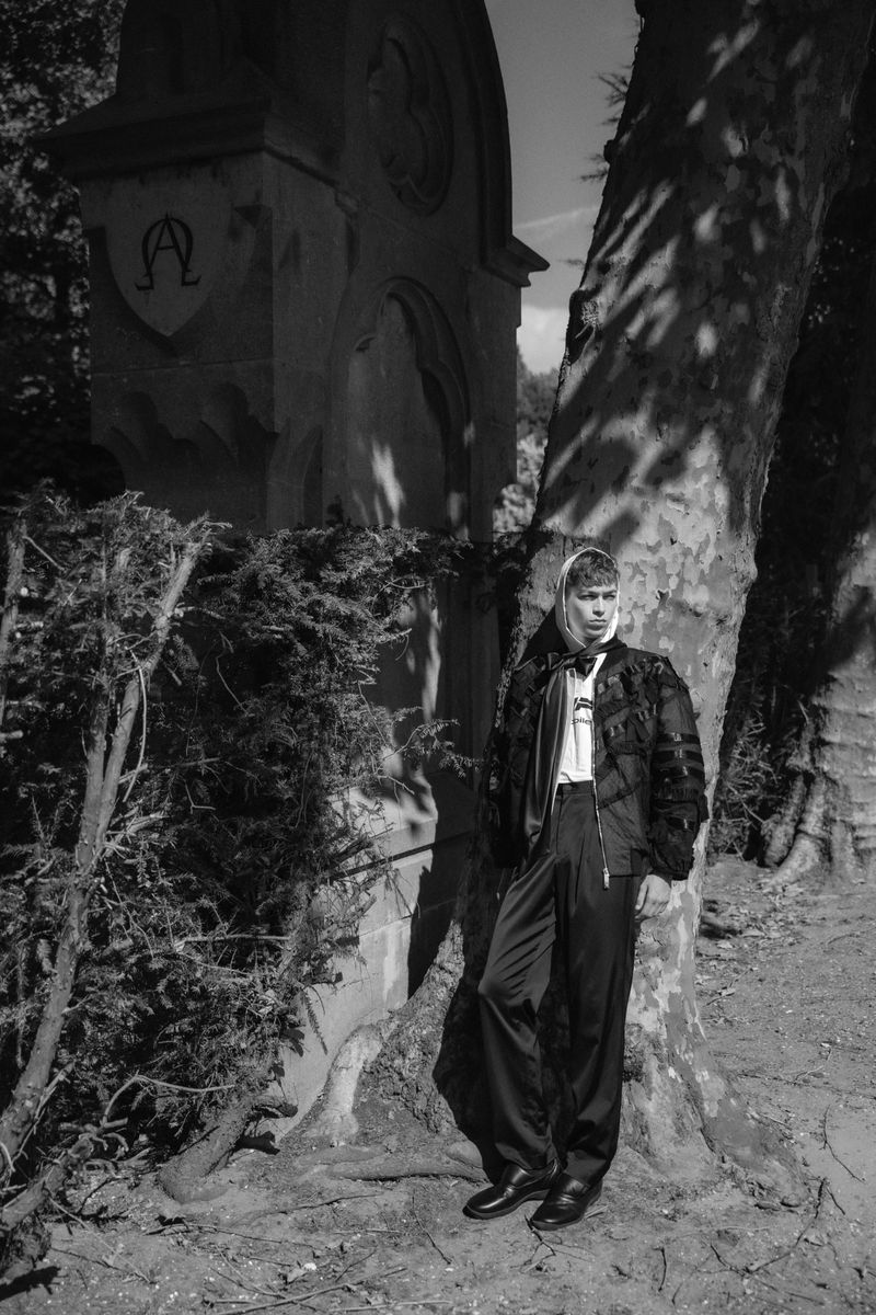 Model standing outdoors near an architectural monument and large tree in a black and white fashion editorial.