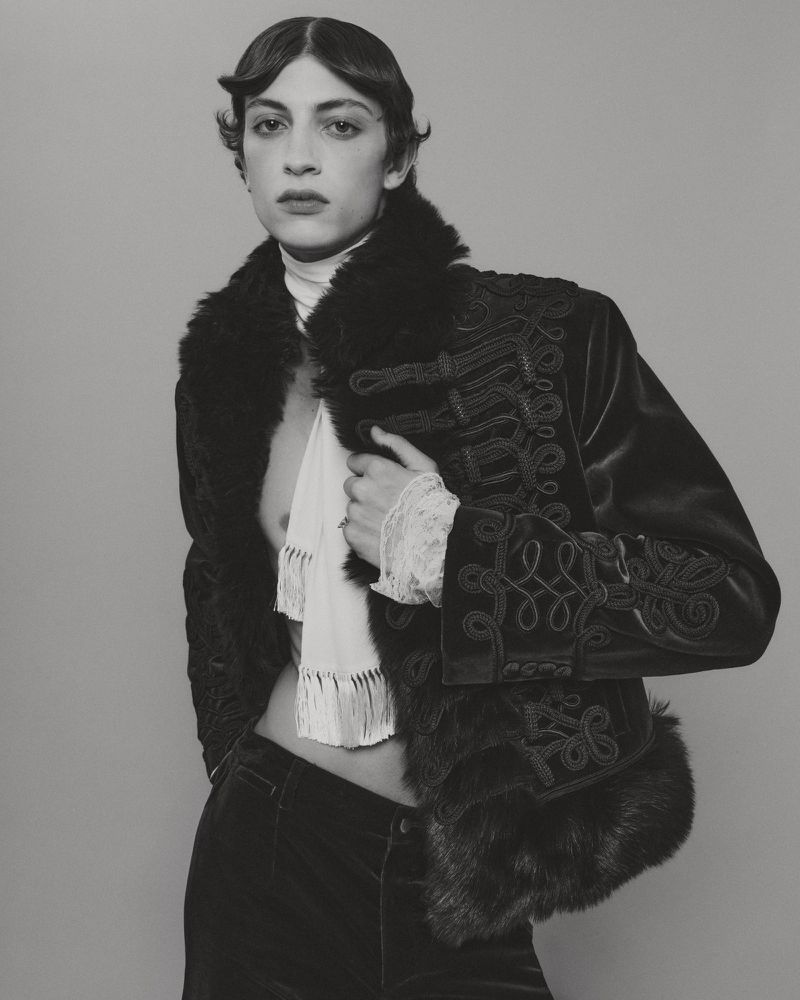 Model in an ornate embroidered velvet jacket with faux fur trim and a white scarf against a seamless grey backdrop.