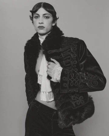 Model in an ornate embroidered velvet jacket with faux fur trim and a white scarf against a seamless grey backdrop.