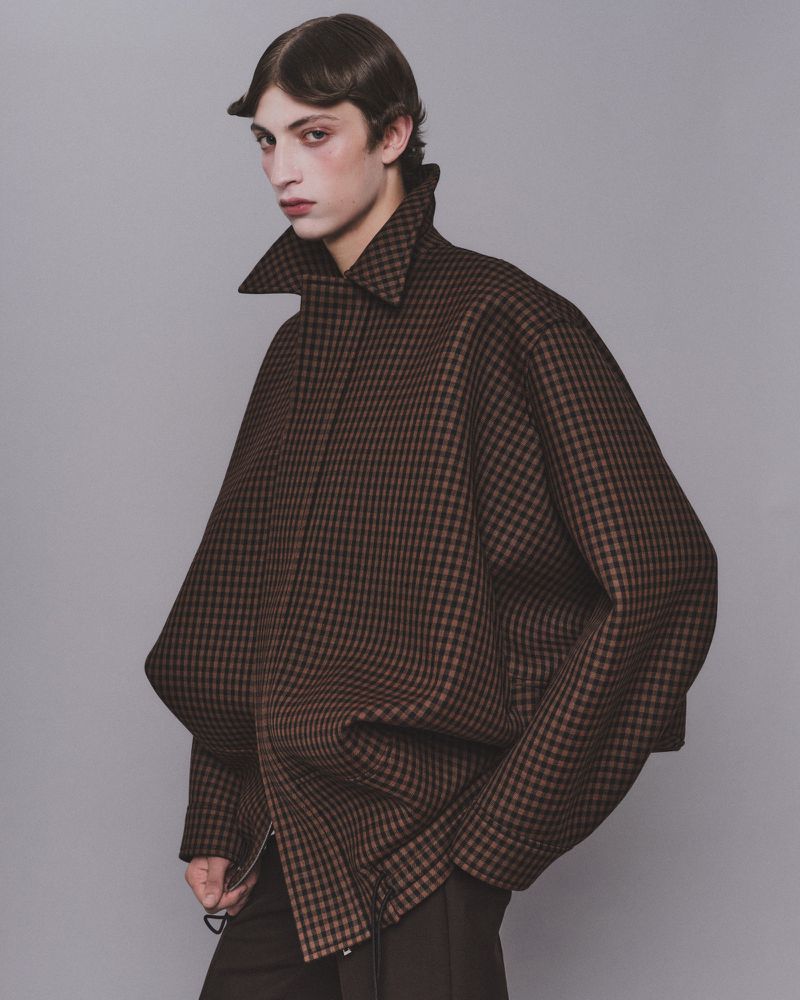 Model wearing a structured brown checkered coat with a high collar against a grey background