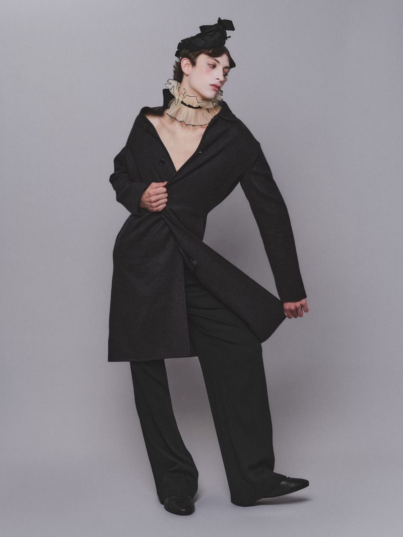 Model in black coat wearing a ruffled collar and black headpiece against a grey background