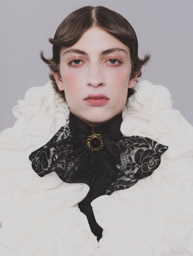Portrait of a model wearing a ruffled white collar and a black lace cravat with a gold brooch against a plain grey background