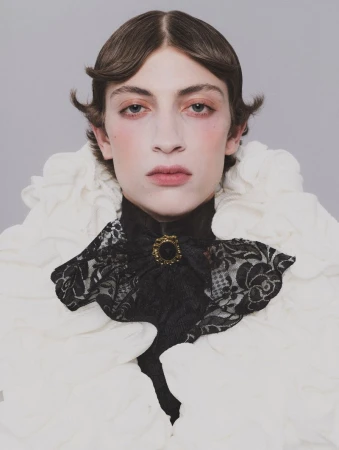 Portrait of a model wearing a ruffled white collar and a black lace cravat with a gold brooch against a plain grey background