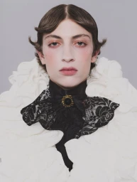 Portrait of a model wearing a ruffled white collar and a black lace cravat with a gold brooch against a plain grey background