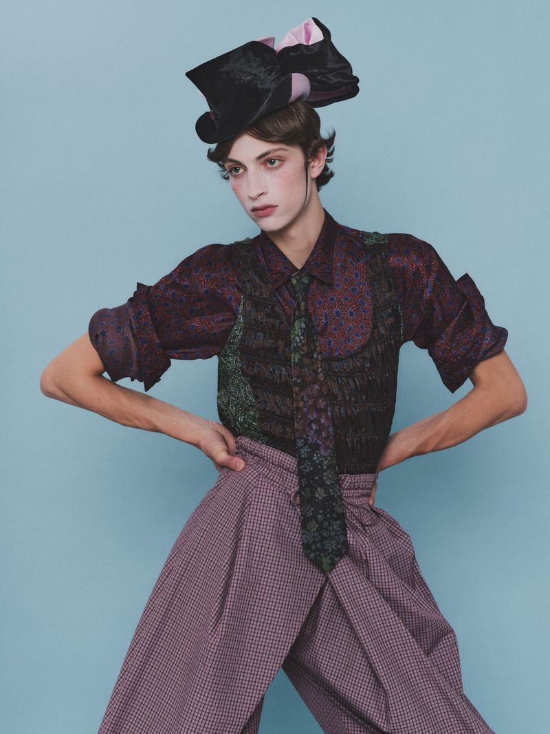 Model Amedeo Mancini in patterned shirt and trousers wearing a black hat with pink bows against blue background for HERO Magazine