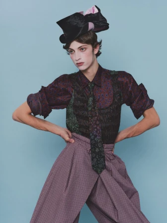 Model Amedeo Mancini in patterned shirt and trousers wearing a black hat with pink bows against blue background for HERO Magazine