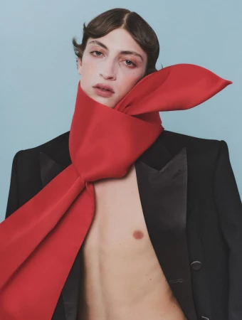 Male model wearing a black blazer and an oversized red silk scarf tied around his neck against a plain pale blue background