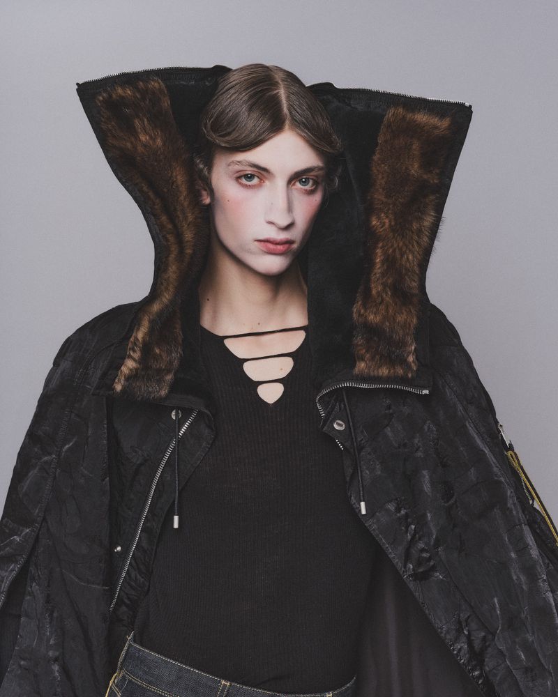 Model wearing a black hooded jacket with brown faux fur trim and a cut-out black top against a grey background
