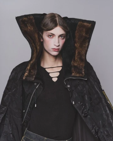 Model wearing a black hooded jacket with brown faux fur trim and a cut-out black top against a grey background