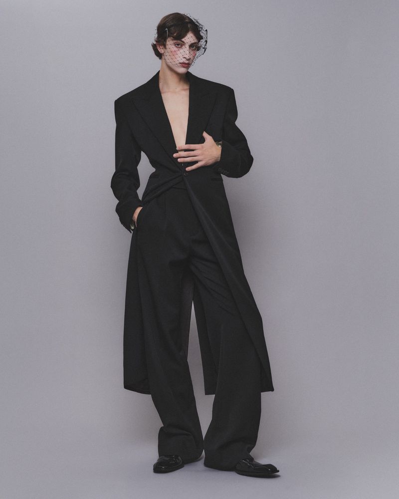Model wearing a black tailored coat and trousers with a netted face veil against a grey background.