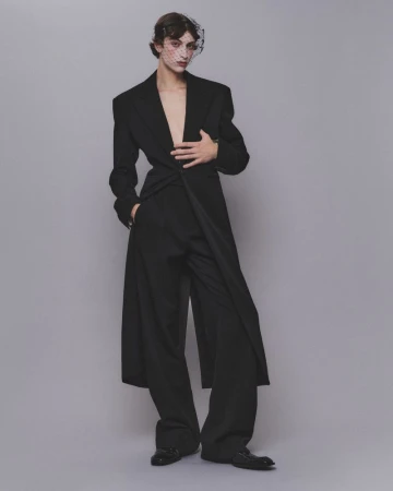 Model wearing a black tailored coat and trousers with a netted face veil against a grey background.