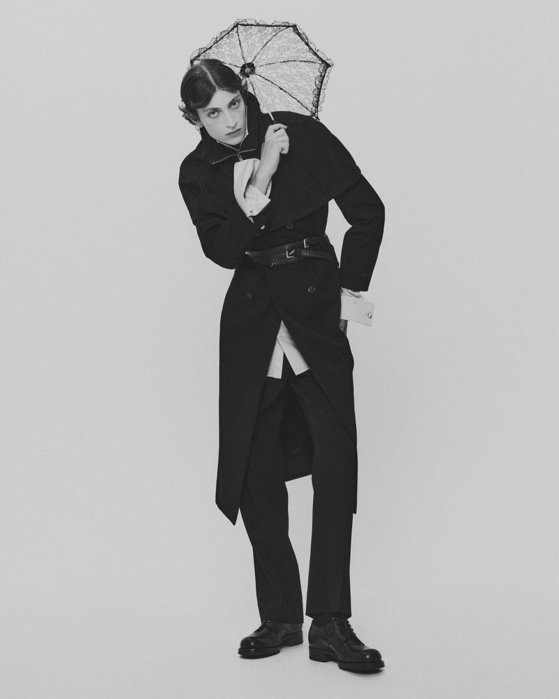 Model wearing a dark long coat and holding a small lace parasol against a plain light studio background.