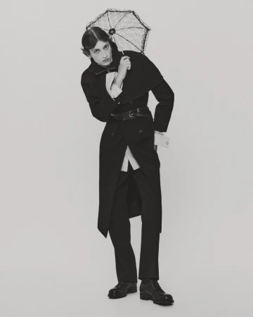 Model wearing a dark long coat and holding a small lace parasol against a plain light studio background.