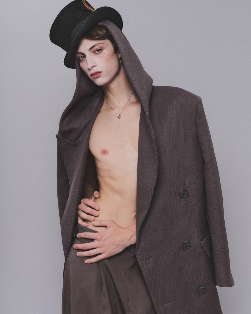 Amedeo Mancini wearing a black top hat and oversized coat against a grey studio backdrop for HERO Magazine.