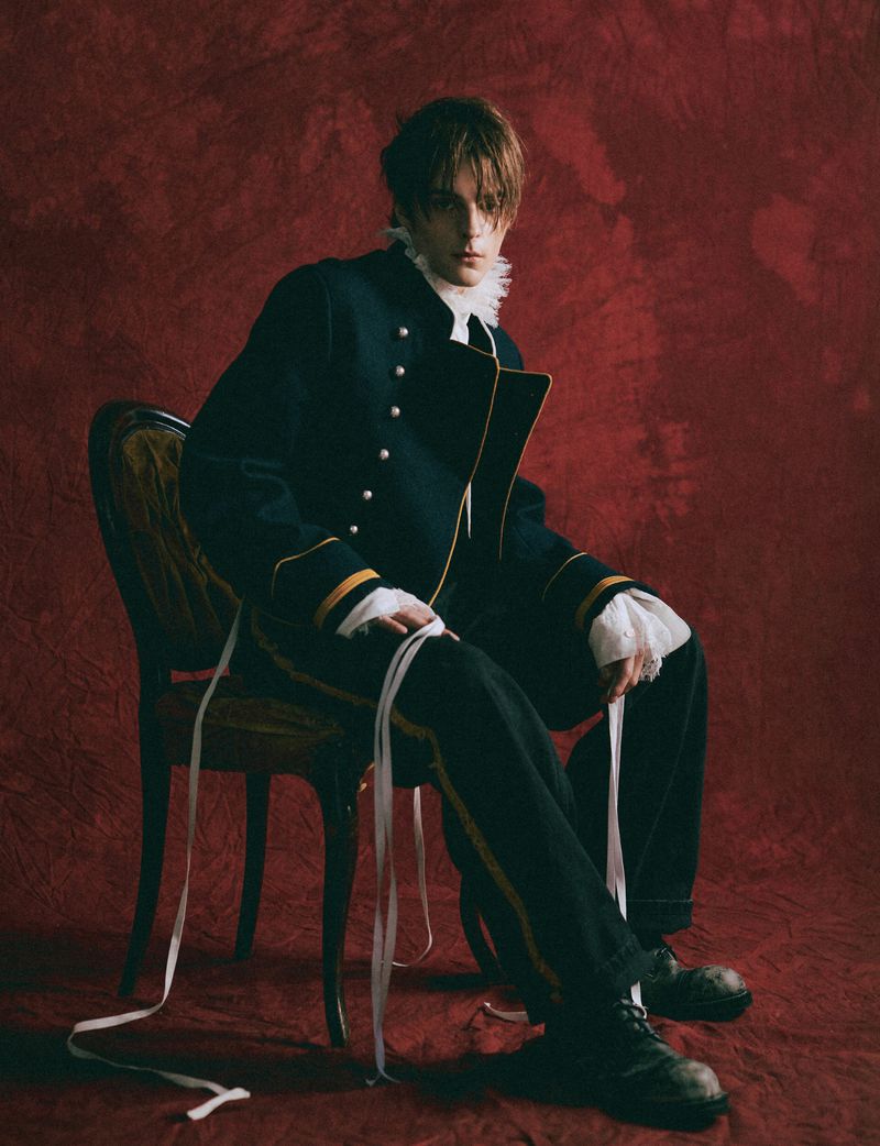 Corey Fogelmanis seated on an antique chair wearing a dark military-style jacket against a textured red backdrop.