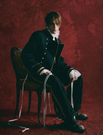 Corey Fogelmanis seated on an antique chair wearing a dark military-style jacket against a textured red backdrop.