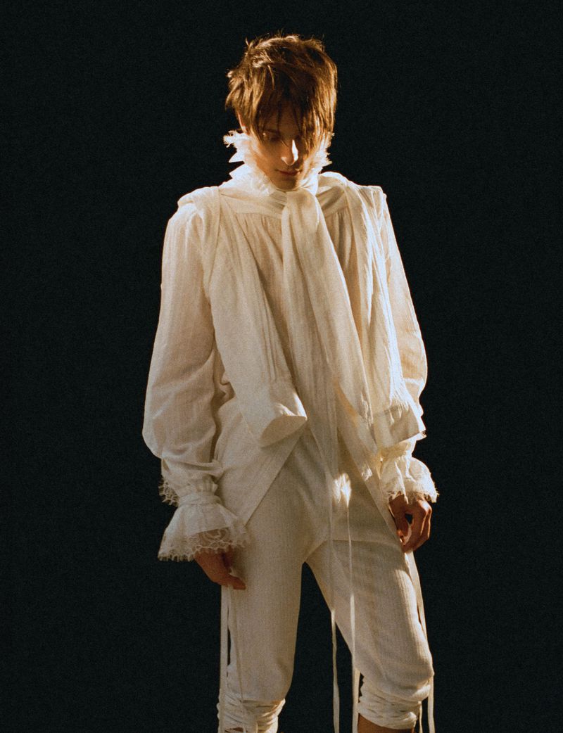 Young man in a white ruffled historical-style shirt and matching trousers standing against a dark background.