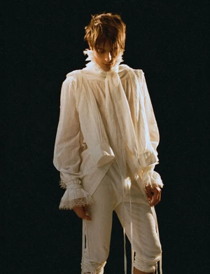 Young man in a white ruffled historical-style shirt and matching trousers standing against a dark background.