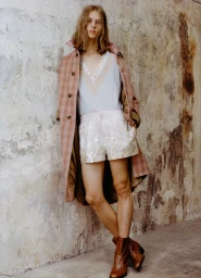 Model with long brown hair leaning against a weathered wall wearing a pink plaid coat, light top, and brown boots.