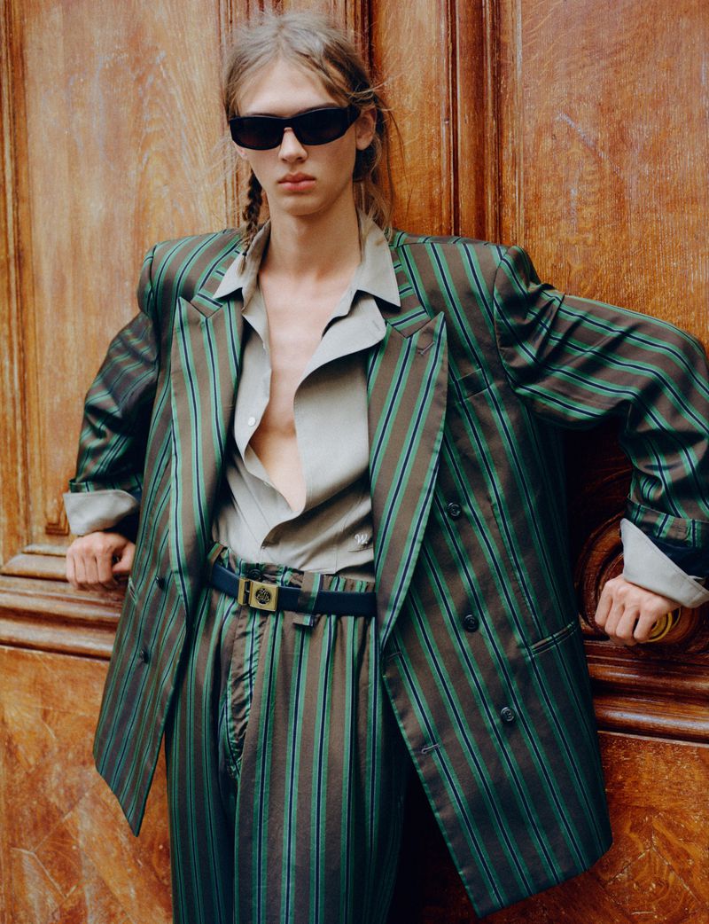 Model in sunglasses wearing a green and brown striped blazer leaning against a wooden door