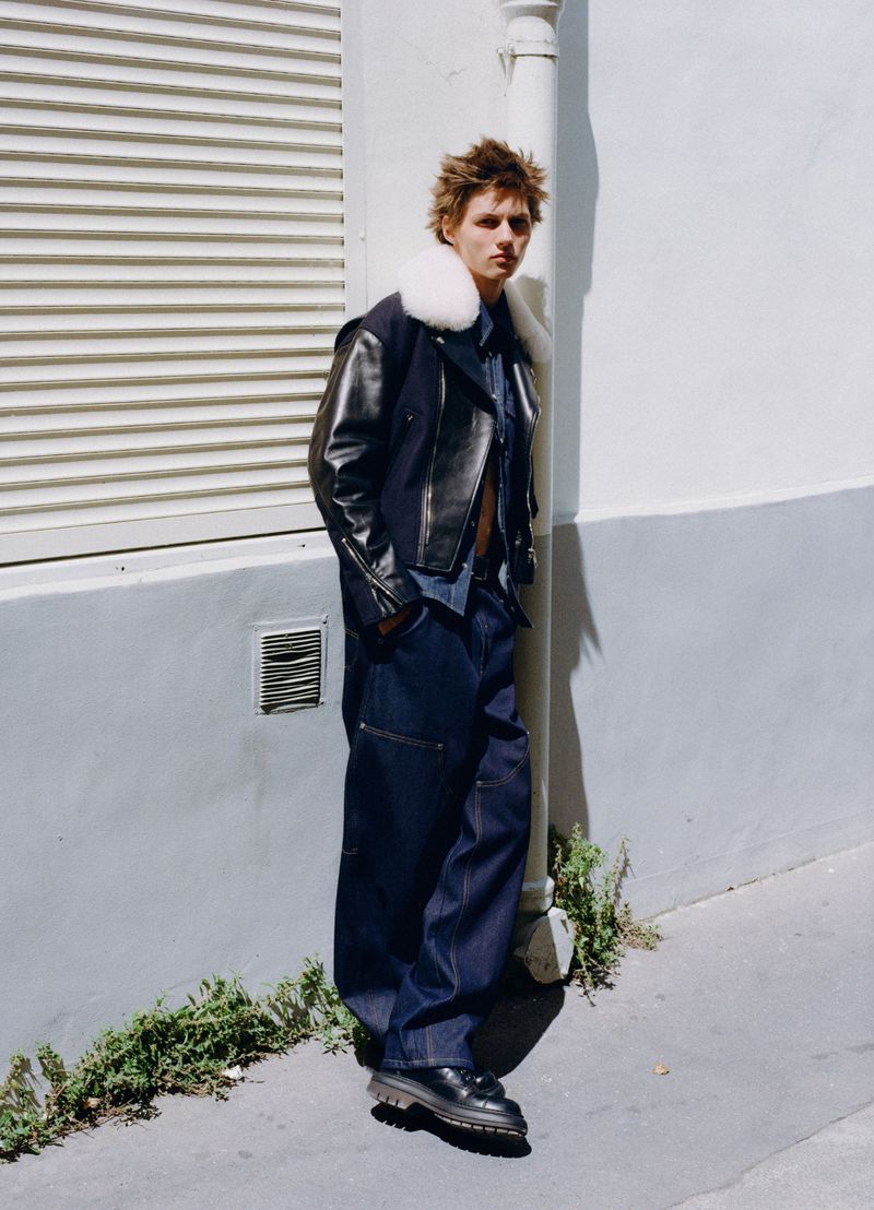 Young male model wearing dark jacket with white collar and wide-leg denim pants leaning against a light wall outdoors.