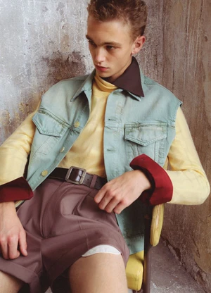 Young man in layered pastel outfit with a light green vest and brown trousers seated against a distressed wall.