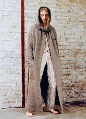Male model in a long brown hooded coat and patterned trousers standing before a weathered brick wall.