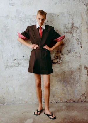 Young male model posing in brown structured shorts and blazer suit against a weathered textured wall