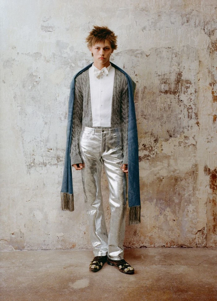 Young male model wearing metallic silver trousers and a cable-knit sweater standing against a weathered concrete wall.
