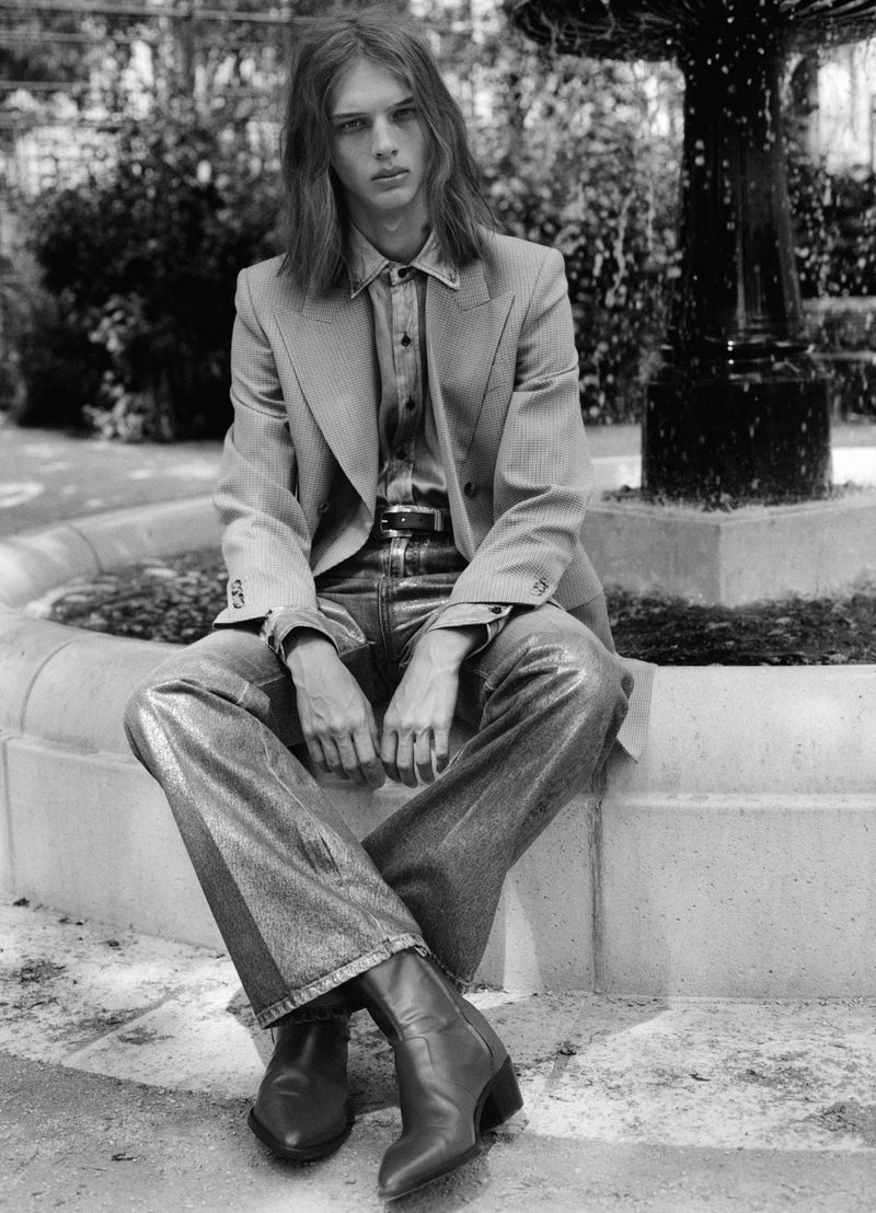 Long-haired male model seated on a stone fountain ledge wearing a blazer and trousers in an outdoor park setting.