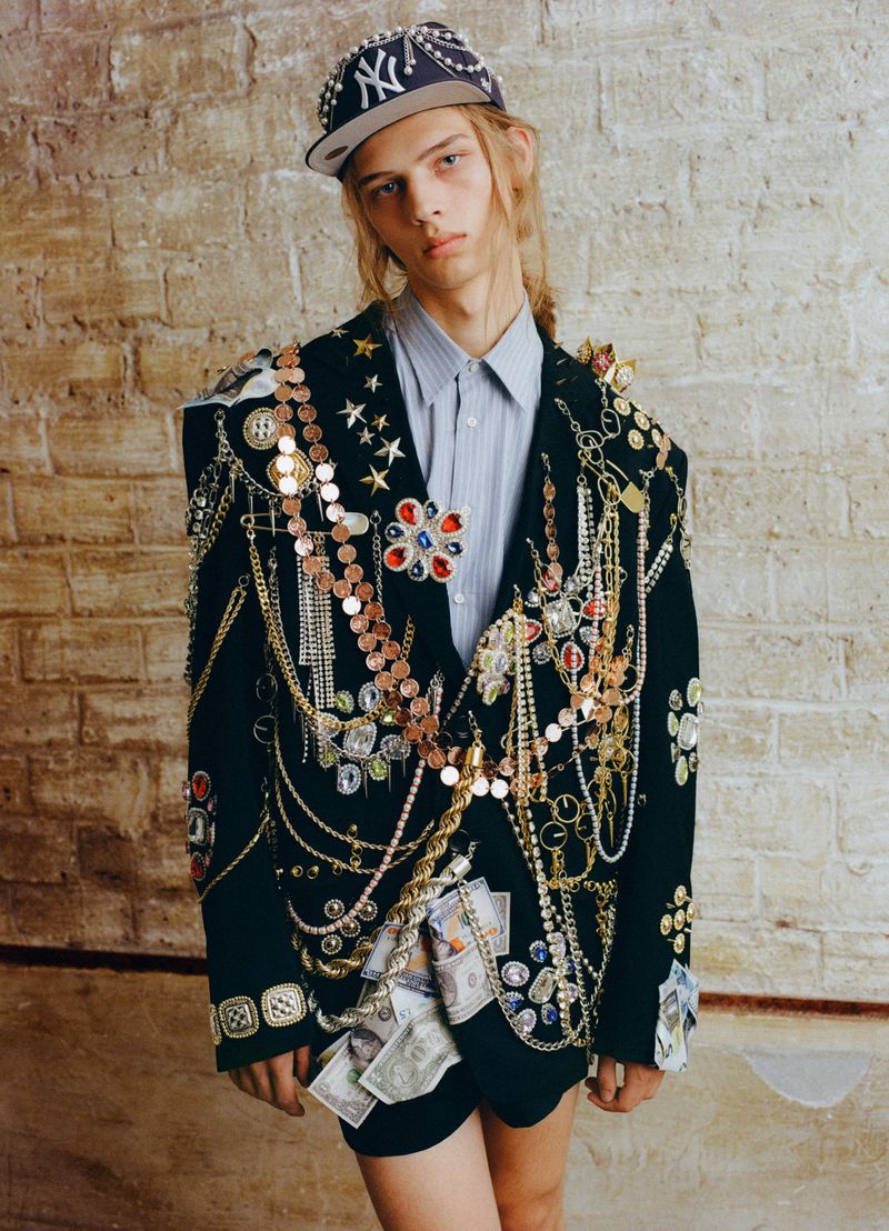 Model wearing heavily embellished black blazer, striped shirt and jeweled baseball cap against textured brick wall