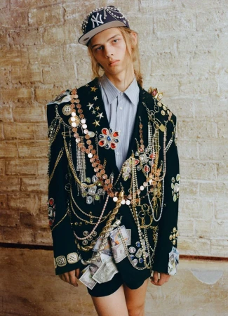 Model wearing heavily embellished black blazer, striped shirt and jeweled baseball cap against textured brick wall