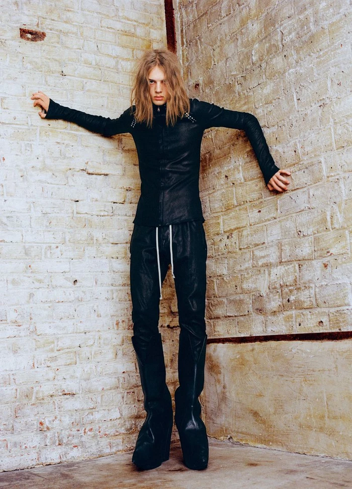 Model with long blonde hair in black jacket and trousers leaning against a white brick corner