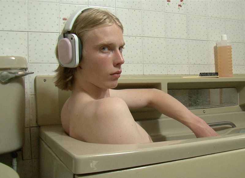 Young man with blond hair wearing white headphones sitting in a bathtub inside a tiled bathroom.