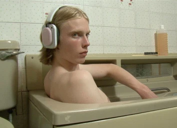 Young man with blond hair wearing white headphones sitting in a bathtub inside a tiled bathroom.