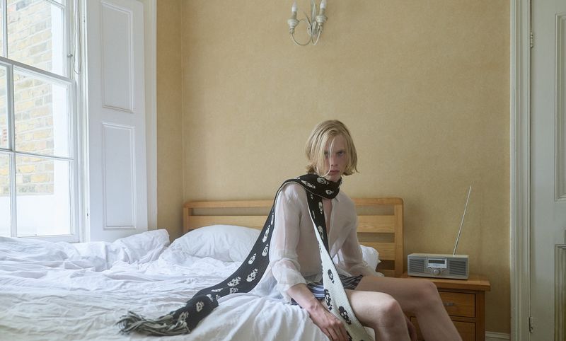 Young man with blonde hair sitting on a bed draped in a long patterned scarf for HERO Magazine Trinkets editorial