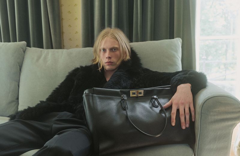 Blonde male model wearing a dark furry jacket and dark trousers, seated on a light grey sofa with a large black leather handbag.