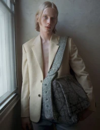 A young male model with long blonde hair wearing a beige blazer and carrying a textured grey shoulder bag indoors near a window.