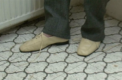 Close-up of cream leather lace-up shoes and grey trousers standing on patterned tiled flooring near a white radiator.