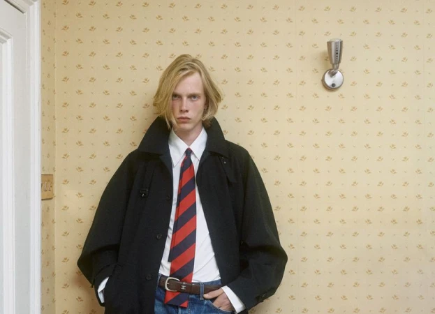 Young male model with blond hair wearing a black coat, white shirt, and striped tie against vintage wallpaper